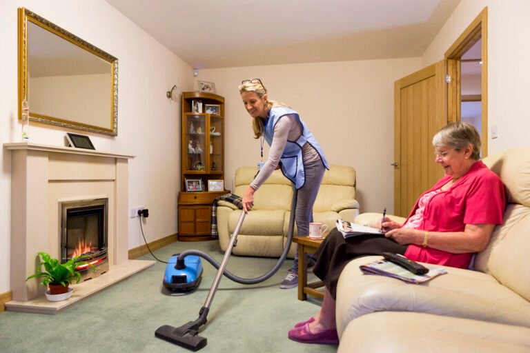 Care worker making a home visit. Female carer is hoovering the living room to help an elderly woman. The woman is sitting on a sofa relaxing